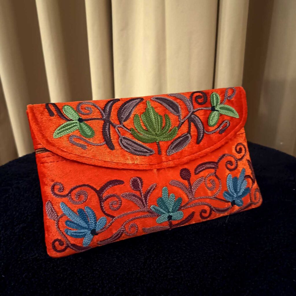 The orange envelope clutch square to the camera on black, flap closed, the embroidered lotus and fan-flower field filling the whole face