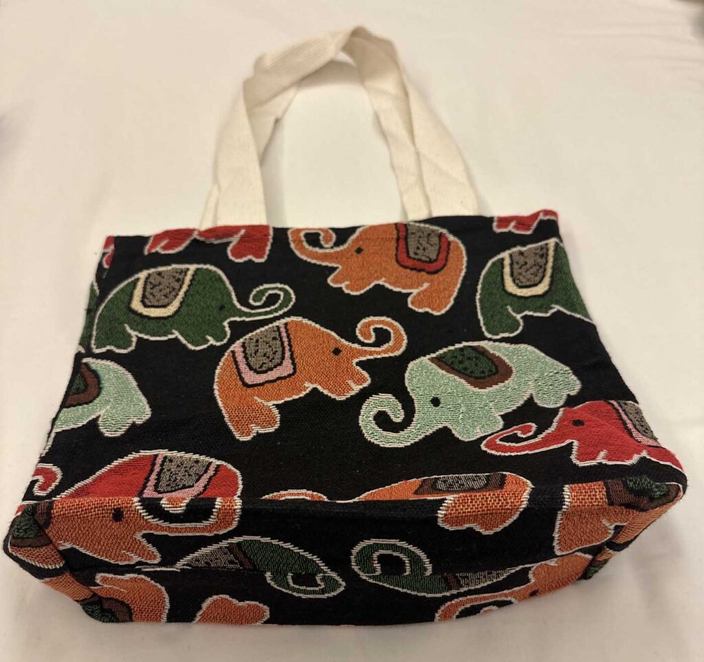 Black woven tote with cream handles, patterned with elephants in olive green, terracotta, mint and red, each outlined in cream thread