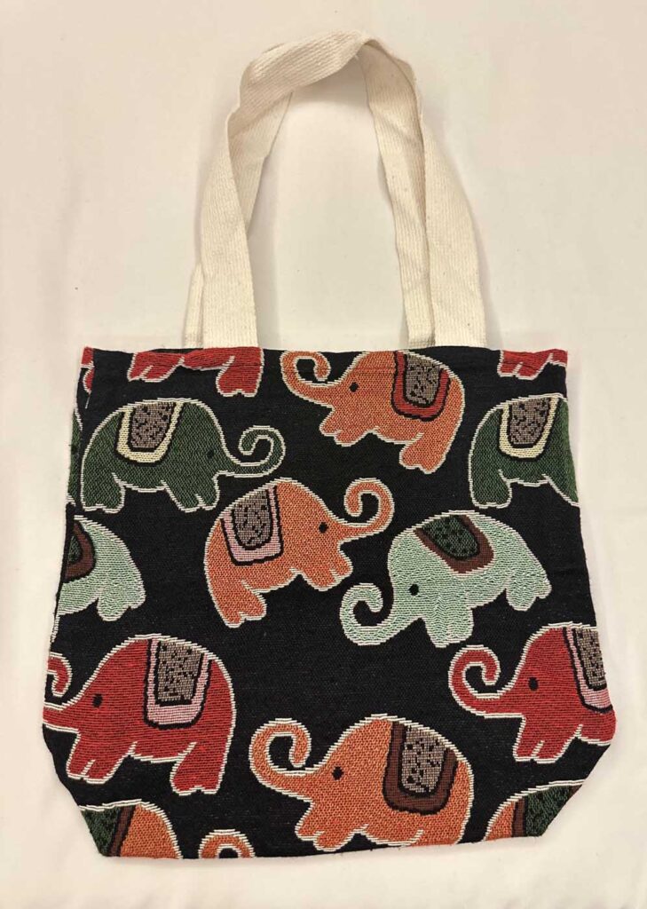 The black elephant tote laid flat with its cream handles extended upward, the olive, terracotta and mint elephants filling the front panel above a horizontal fold
