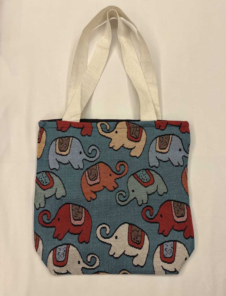 Teal-blue woven tote with cream handles, patterned with elephants in red, periwinkle, orange, pale green and white on a blue-green ground