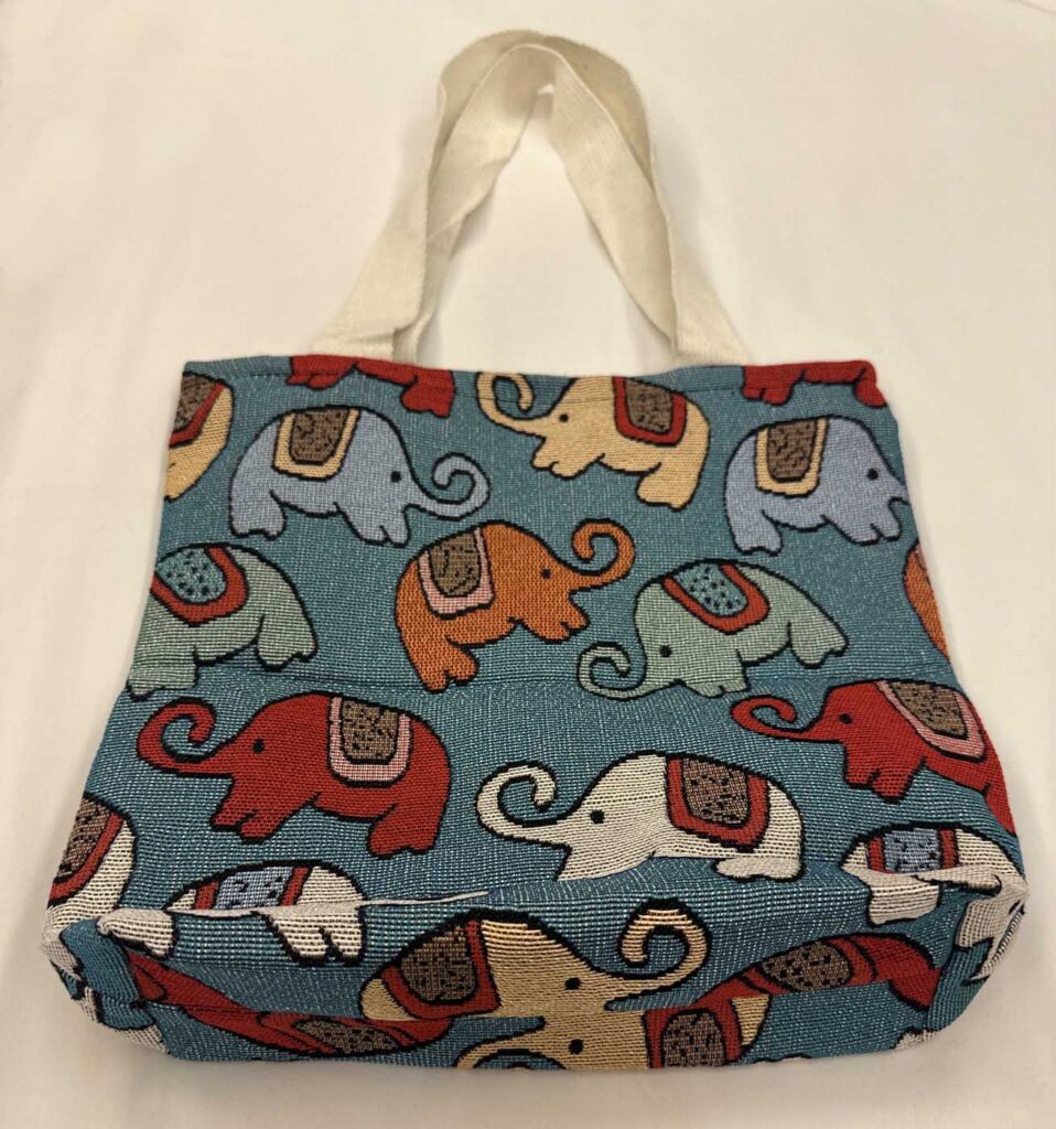 The teal-blue tote seen from a slight angle so the base panel and its continuing elephant pattern are visible