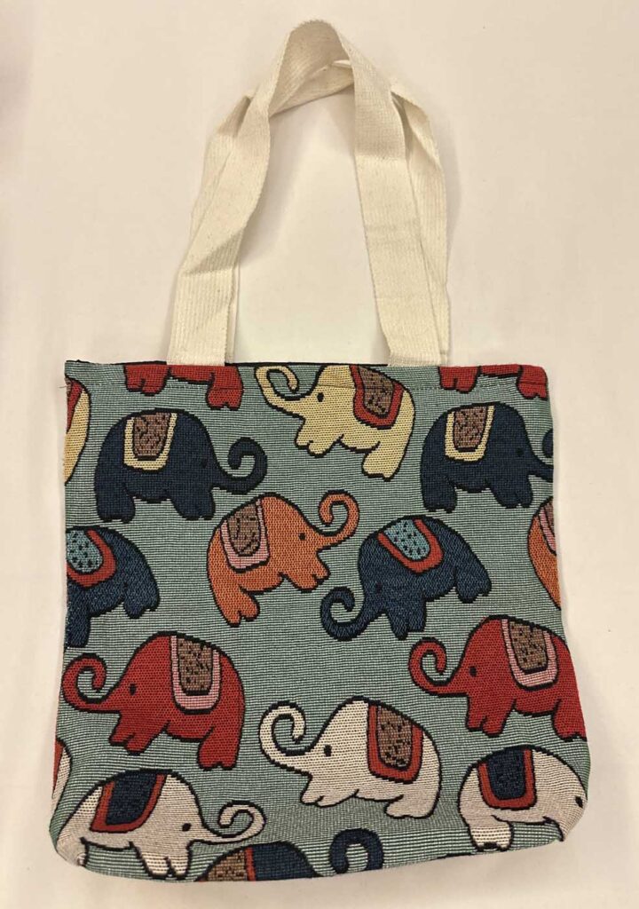 Sage-green woven tote with cream handles, patterned with elephants in navy, red, orange, pale blue and cream, each with a woven saddle blanket