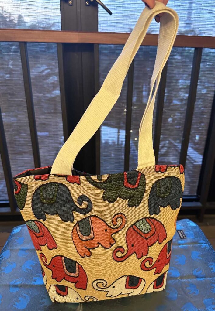 Cream-yellow elephant tote hanging by its long shoulder strap from a door handle, on a balcony with bamboo blinds behind it; elephants in charcoal, orange, red and white