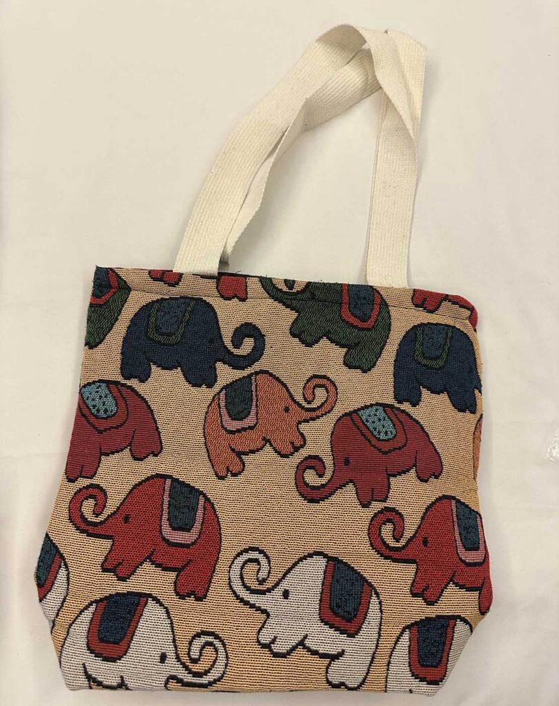 Front view of the cream-yellow elephant tote on a pale ground, handles laid back, elephants in navy, red, orange and white across a warm sand-coloured weave