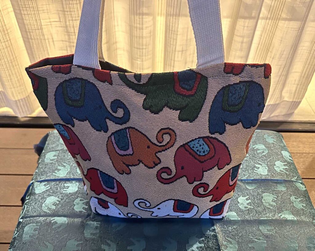 The cream-yellow tote standing upright on a printed cloth, lit warmly so the ground reads taupe and the elephants navy, dark green, red and orange