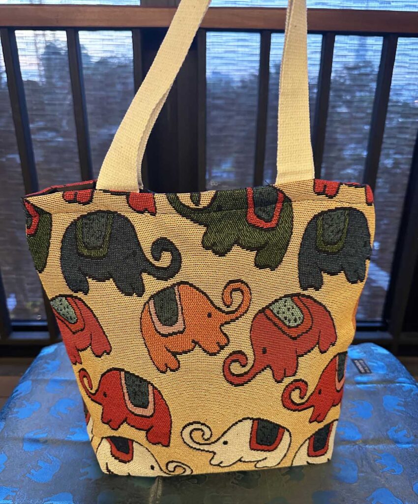 The cream-yellow elephant tote standing on a blue elephant-printed cloth, its long cream strap rising out of frame, charcoal and terracotta elephants on a sand ground