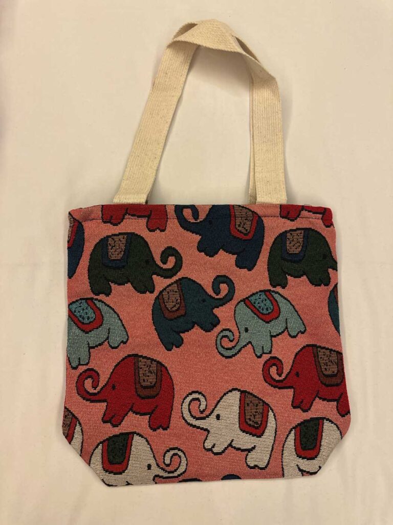 Salmon-pink woven tote with cream handles, patterned with elephants in navy, dark green, teal, red and white, each with a small woven saddle