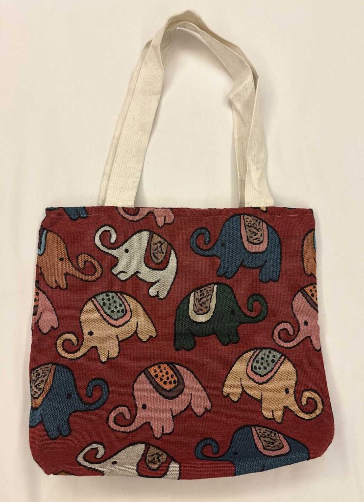 Brick-red woven tote bag with cream canvas handles, covered in a repeating pattern of elephants in blue, pink, cream, olive and tan, each wearing a patterned saddle blanket