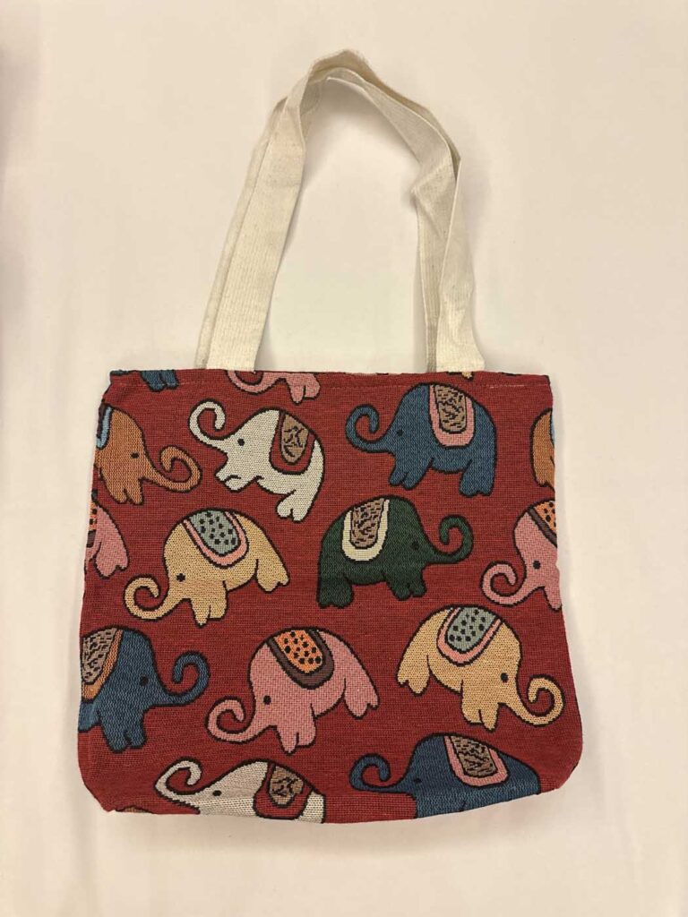 Front view of the brick-red elephant tote laid flat, its cream handles folded above it and the woven elephant pattern filling the whole panel