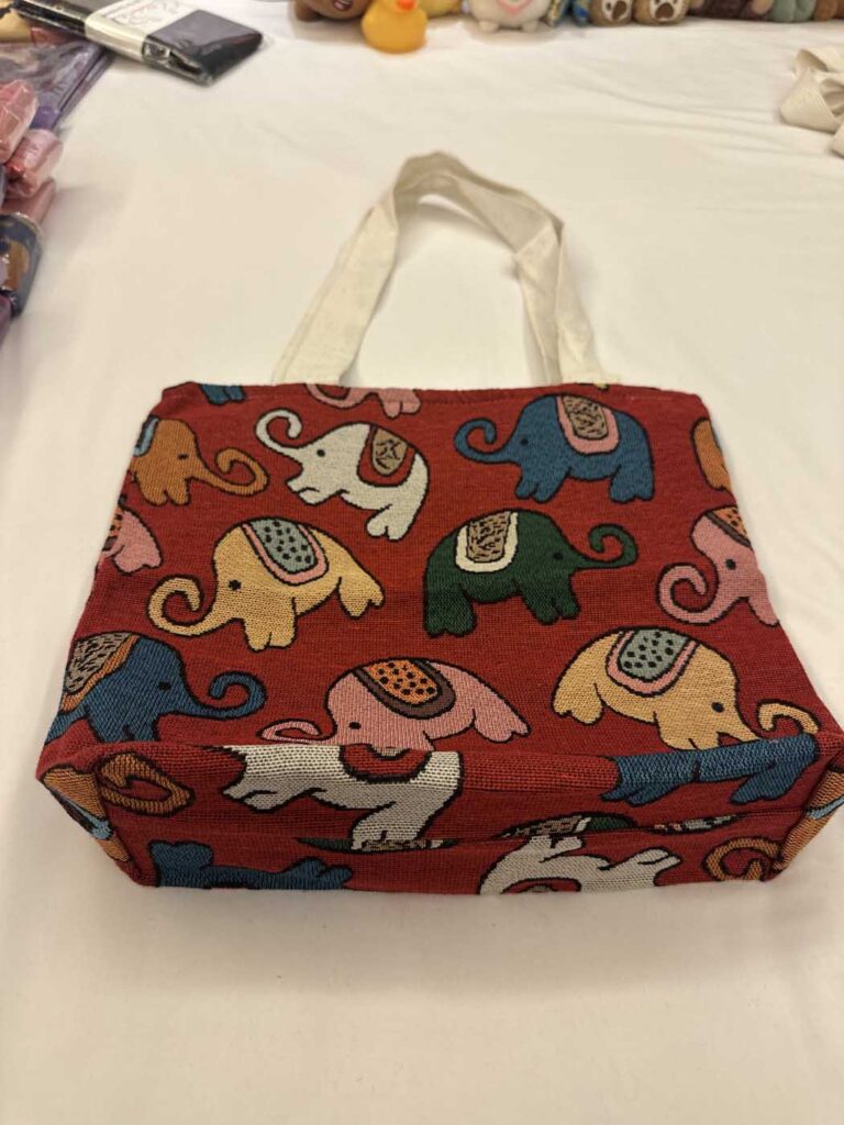 The red elephant tote photographed at an angle on a white bedspread, showing the gusseted base and how the pattern continues around the side