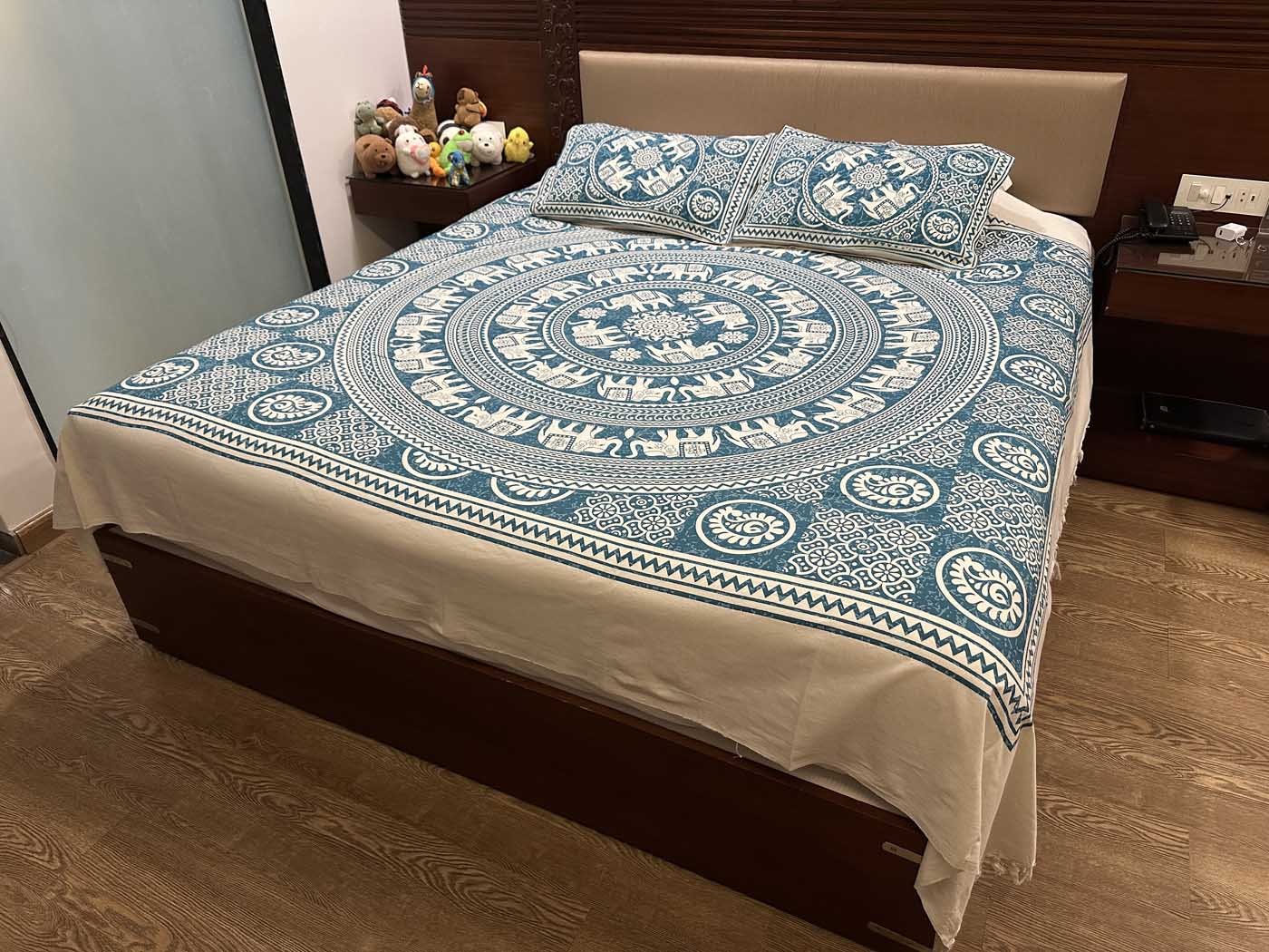 A double bed made up with a white cotton sheet block-printed in turquoise: a large central mandala ringed by rows of elephants, zigzag and paisley borders, and two matching pillow covers against a padded headboard