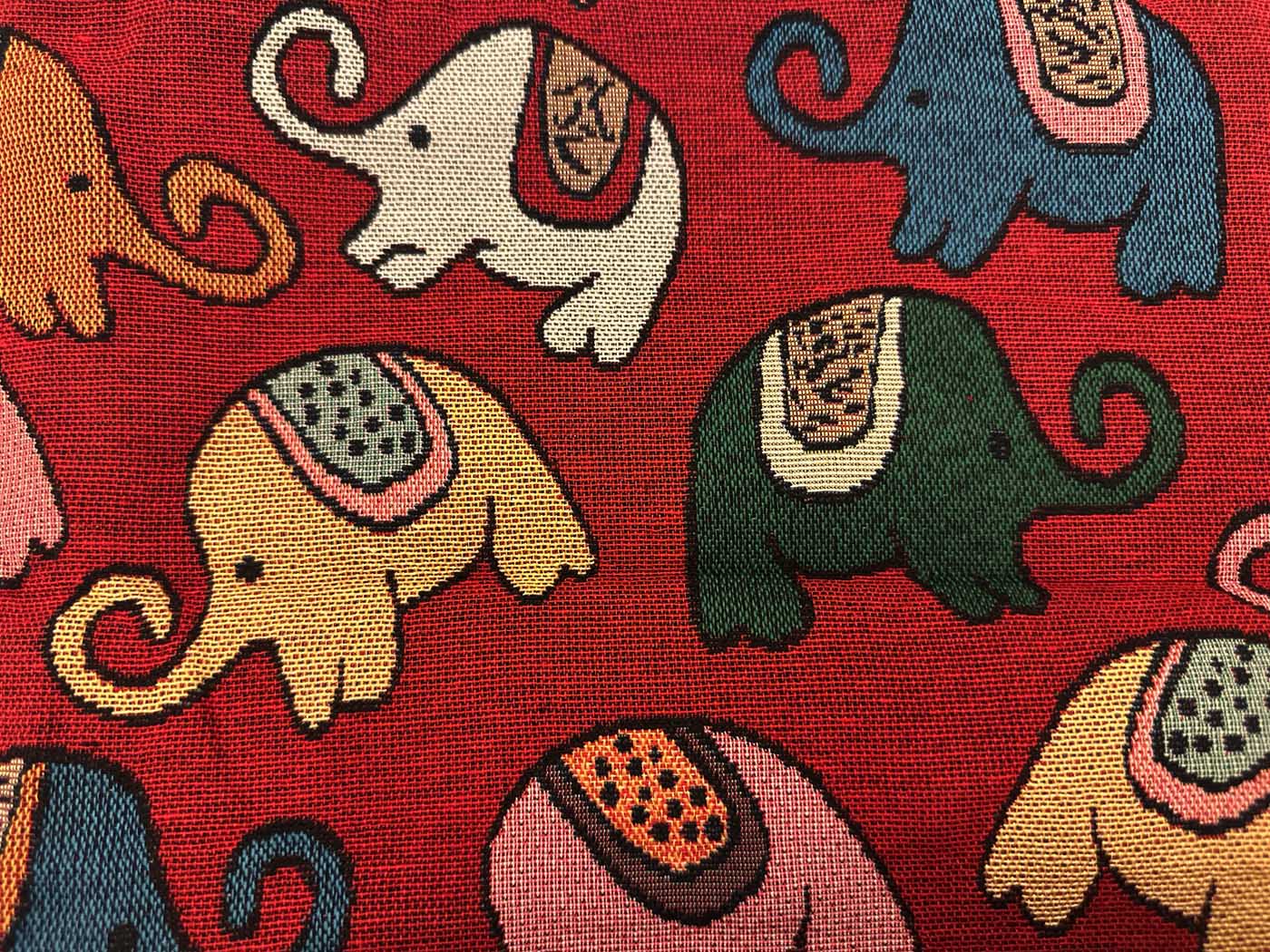 Close view of brick-red woven fabric patterned with elephants in cream, blue, olive, tan and pink, each wearing a small patterned saddle blanket