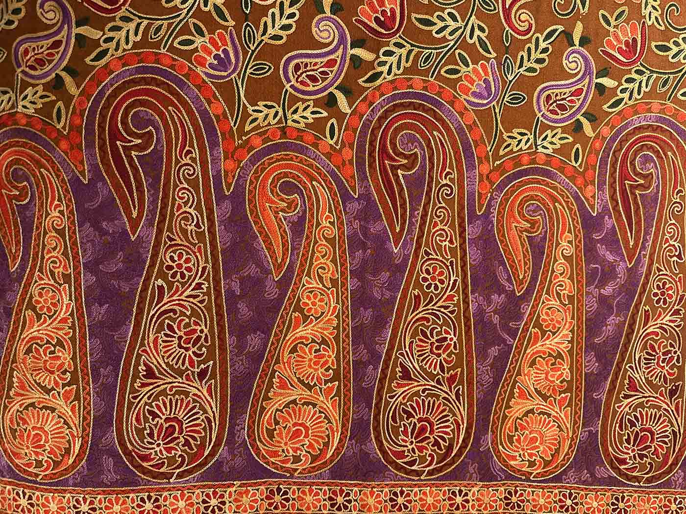 Close view of a tobacco-brown Kashmiri wool stole embroidered with large gold and purple paisleys over a purple ground, edged with chains of coral flowers