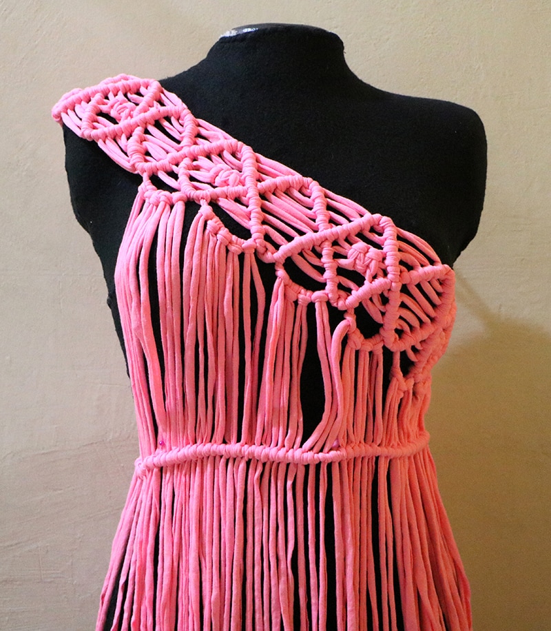 Coral-pink macramé dress on a black dress form, one shoulder worked in a knotted diamond lattice and the skirt falling as loose cords from a knotted waistband