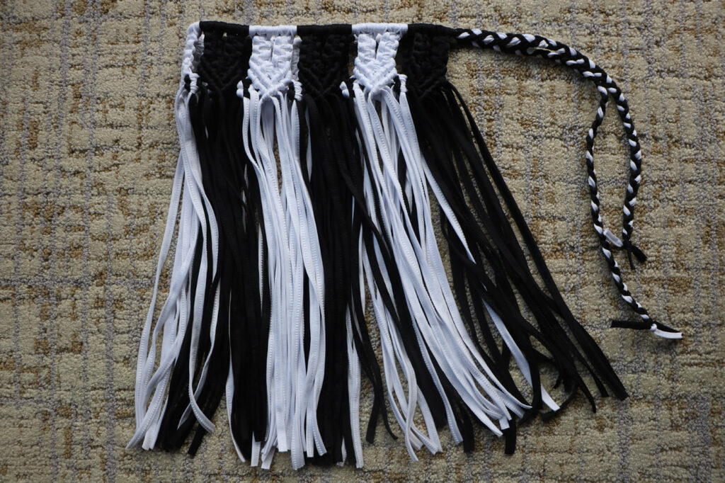 Black-and-white macramé mini skirt laid flat on carpet, alternating panels of knotted black and white cord over a long fringe, a braided two-tone tie extending to one side