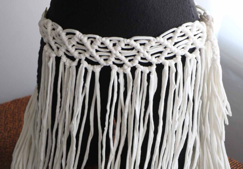 Detail of the long white macramé skirt at the waist: two deep bands of knotwork on a black dress form, with loose cotton cord falling free below them