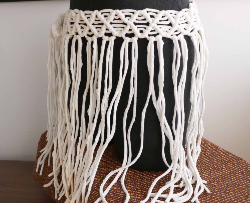 Close view of a white macramé skirt on a black dress form — a waistband of knotted diamond lattice above a long fringe of soft cotton cord, resting on a rust-coloured woven stool