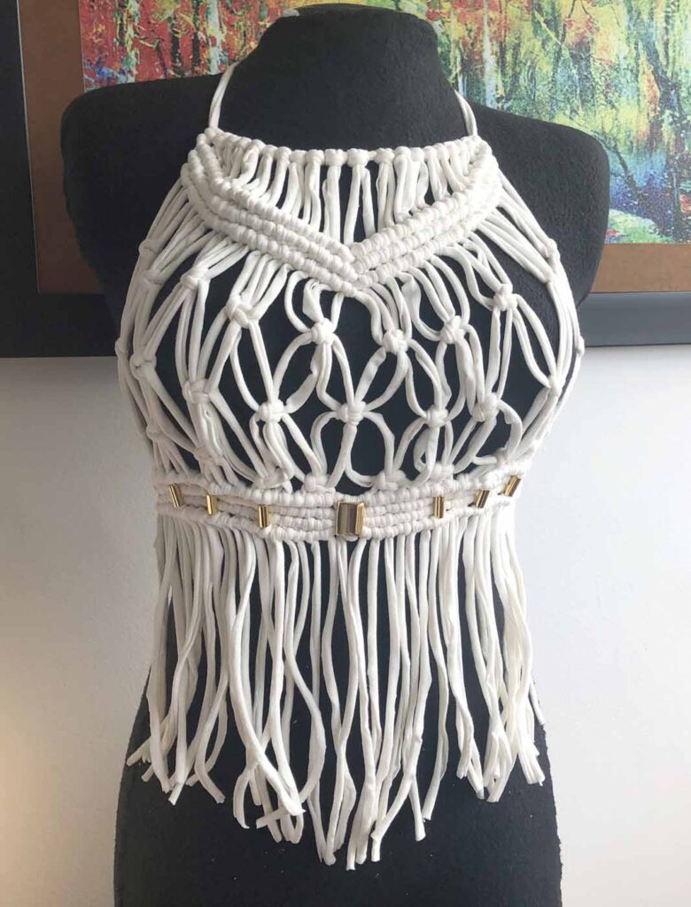 White macramé halter blouse on a black dress form, the bodice knotted in an open flower lattice above a banded waist set with small gold beads and a long cord fringe