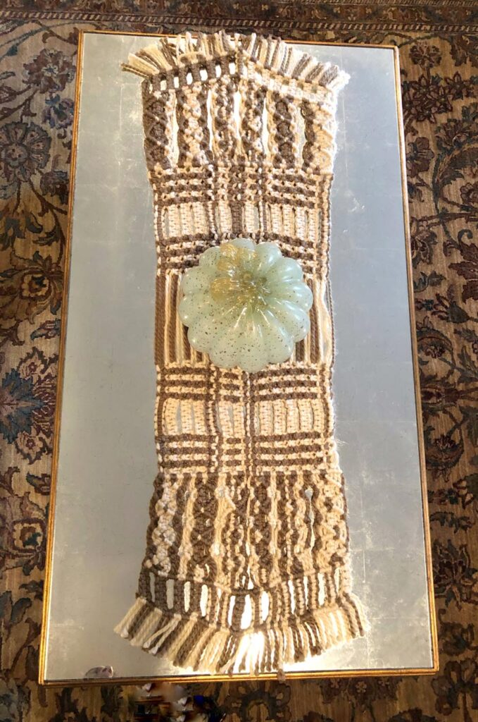 Brown-and-beige macramé panel laid on a gilt-framed mirror, bands of knotwork fringed at both ends with a pale green glass flower dish resting at its centre