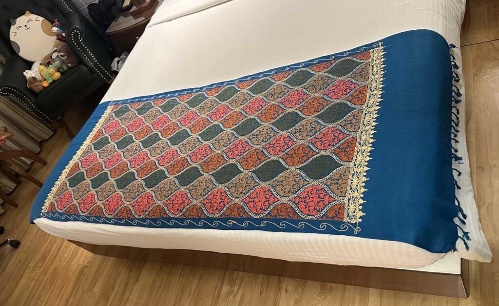 A petroleum-blue Kashmiri wool stole spread diagonally across a white bed, its embroidered end panel of teardrop medallions in coral, orange, olive and gold facing the camera, plain blue body and knotted tassels beyond