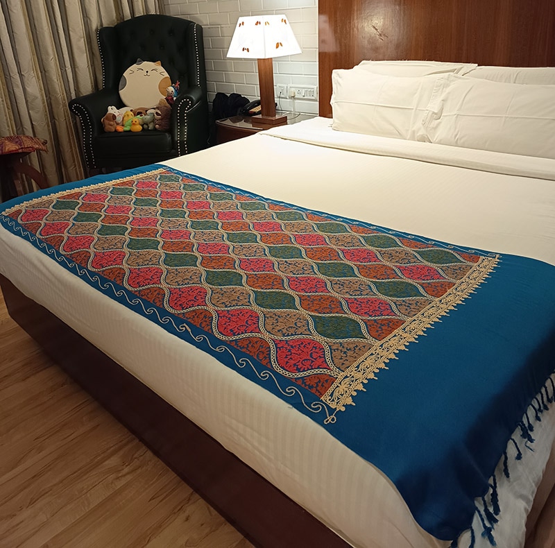 The petroleum-blue stole laid across a made bed as a throw, embroidered panel toward the foot, a table lamp and a dark armchair behind it