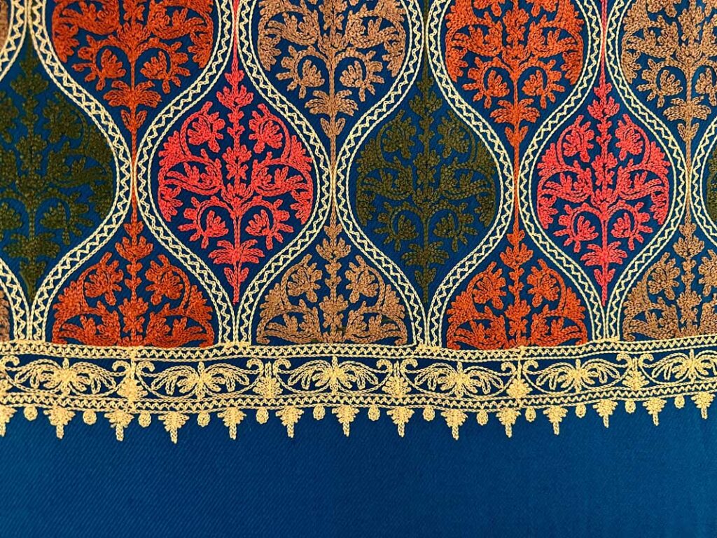 Close view of a deep petroleum-blue Kashmiri wool stole, hand-embroidered with rows of teardrop medallions in coral, burnt orange, olive and gold, finished with a scalloped gold border