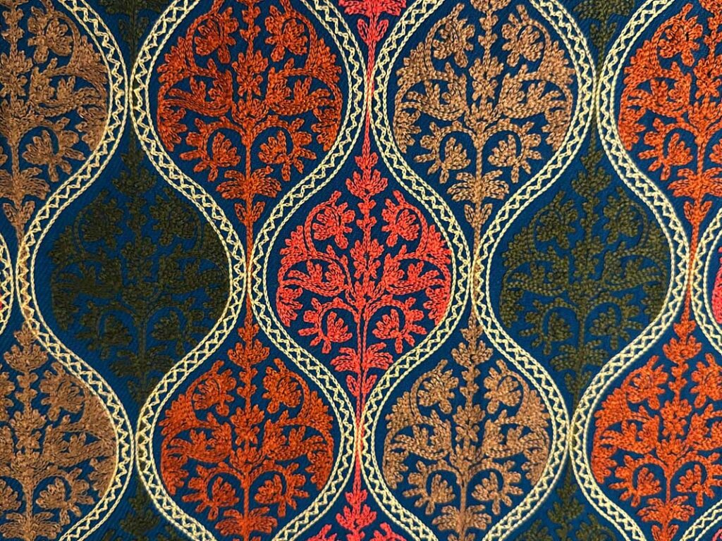 The petroleum-blue field at close range, the cream chain-stitch outlines running in vertical waves and the coral and gold sprays filling each medallion
