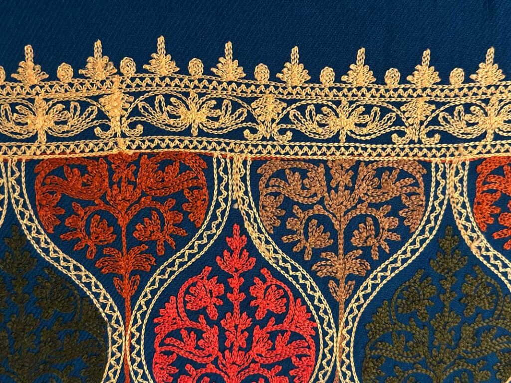 Detail of the gold border band: a row of scrolling cream-gold flowers between narrow braided rules, with a fringe of small embroidered picots above the plain blue wool
