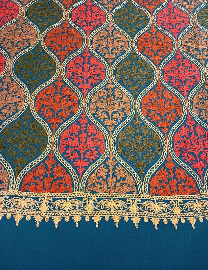 The petroleum-blue stole photographed to show the transition from dense medallion embroidery through the gold border to the plain woven blue hem