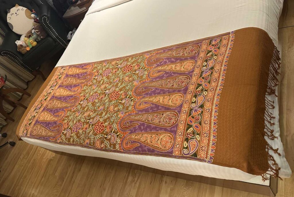 A tobacco-brown Kashmiri wool stole spread on a white bed, its end panel worked in large gold and purple paisleys around a floral centre, plain brown body and knotted tassels at the far end