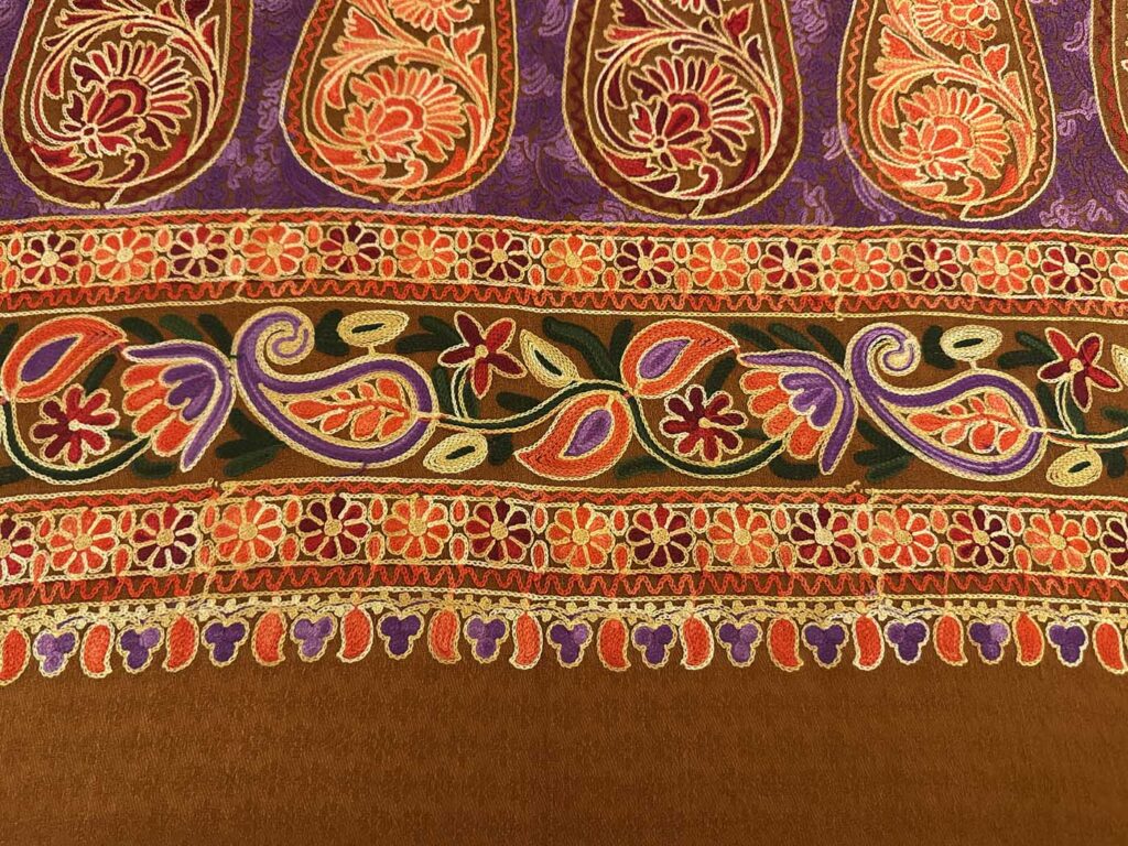 Detail of the brown stole's borders: a row of purple and coral paisleys above two chains of small orange daisies, a scrolling paisley band between them, and a fringe of purple and orange petals over plain brown wool