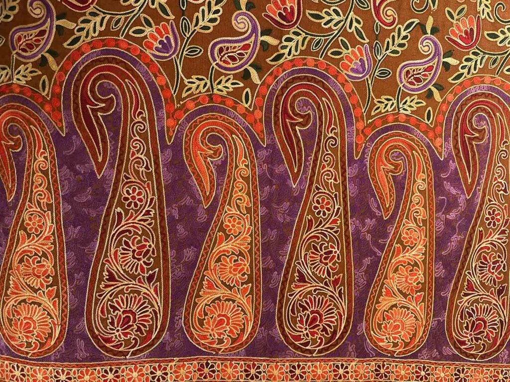 Close view of a tobacco-brown Kashmiri wool stole embroidered with large gold and purple paisleys over a purple ground, edged with chains of coral flowers