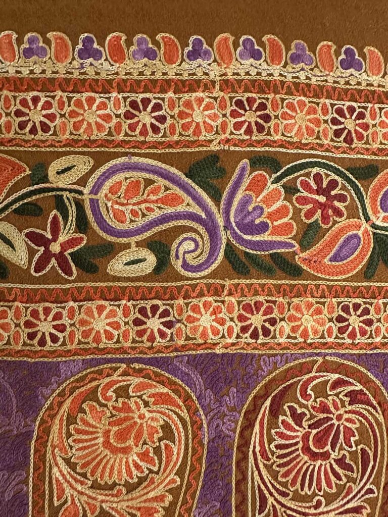 The brown stole's stacked borders at close range, two daisy chains framing a scrolling paisley band, with the purple paisley panel beginning below