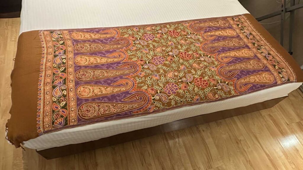 The brown stole seen along the length of a bed, the embroidered paisley panel at the near end and the plain brown body running away toward the tasselled edge
