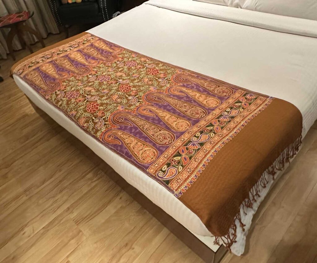 The brown stole laid as a bed throw, showing the full end panel of gold and purple paisleys, its layered floral borders, and the tasselled fringe hanging over the side