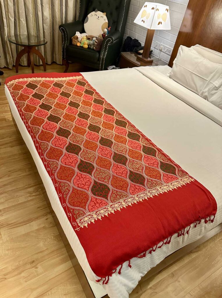 A crimson Kashmiri wool stole laid down the length of a white bed, its embroidered end panel of coral and olive medallions facing the camera, a gold picot edge, and red tassels hanging over the foot