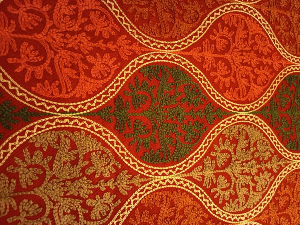 Very close view of the crimson embroidery, the gold chain-stitch waves crossing the frame with orange, olive and tan floral sprays worked between them