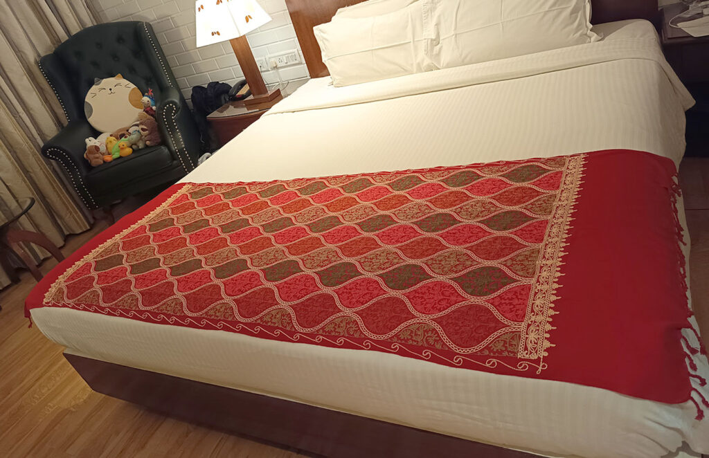 The crimson stole spread across a made bed as a throw, embroidered panel angled toward the camera, a table lamp and dark armchair behind