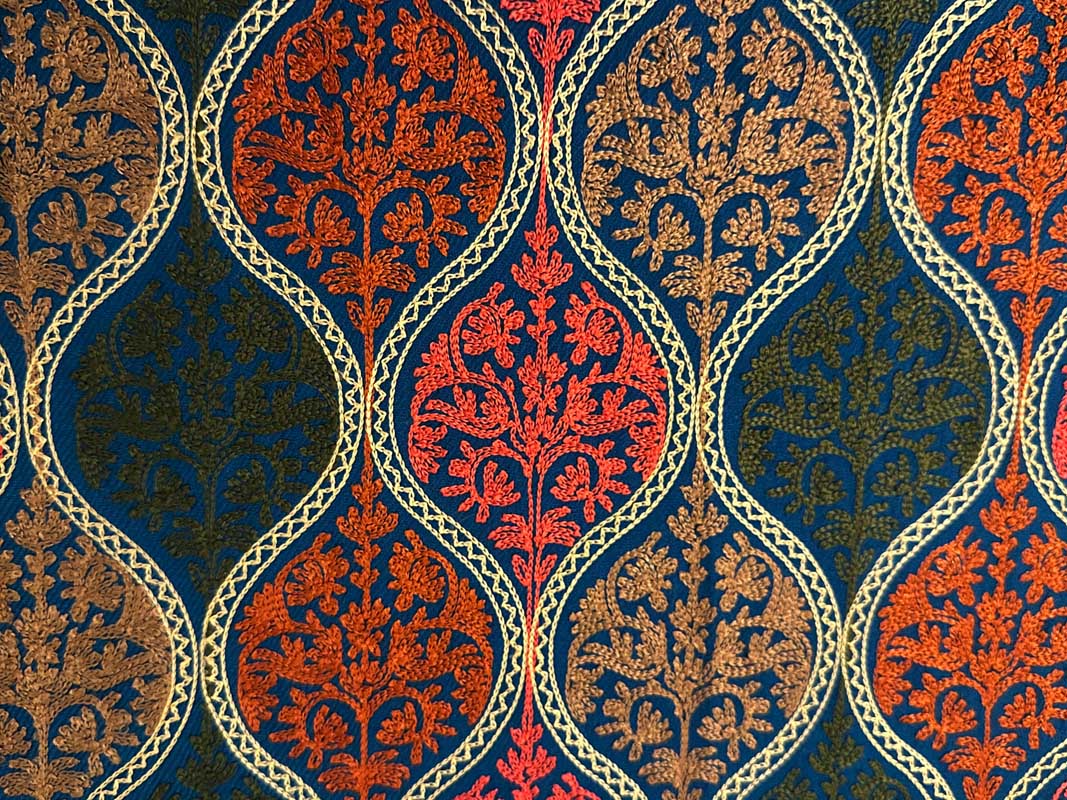 Close view of a petroleum-blue Kashmiri wool stole, its field of teardrop medallions embroidered in coral, burnt orange, tan and olive and outlined in cream chain stitch